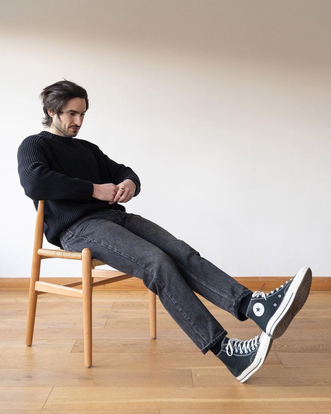 stylish man seated in a chair wearing a black crewneck sweater with black jeans and black high top sneakers