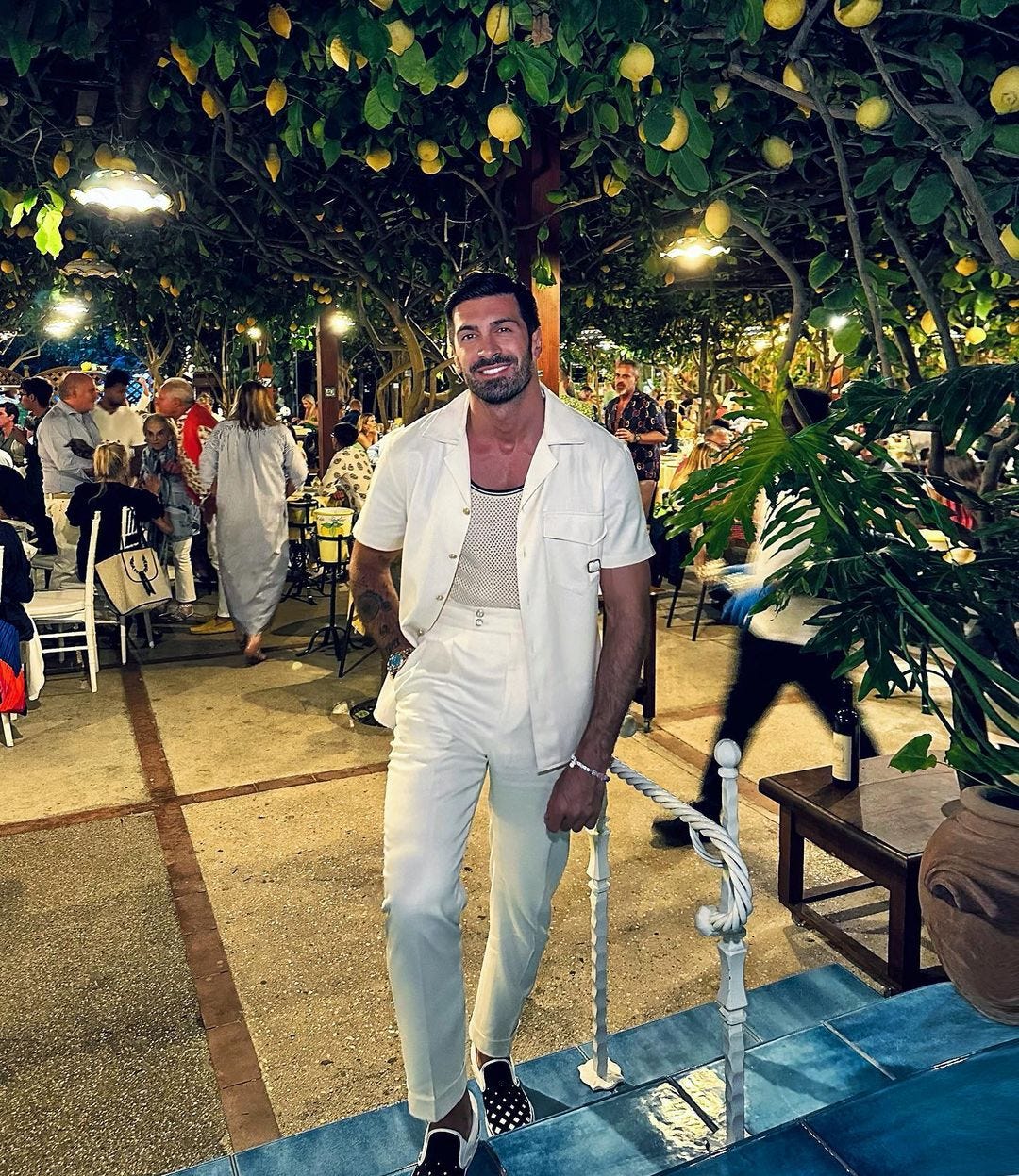 stylish man at a party wearing an off-whtie camp shirt over a tank top with white pants and brown shoes