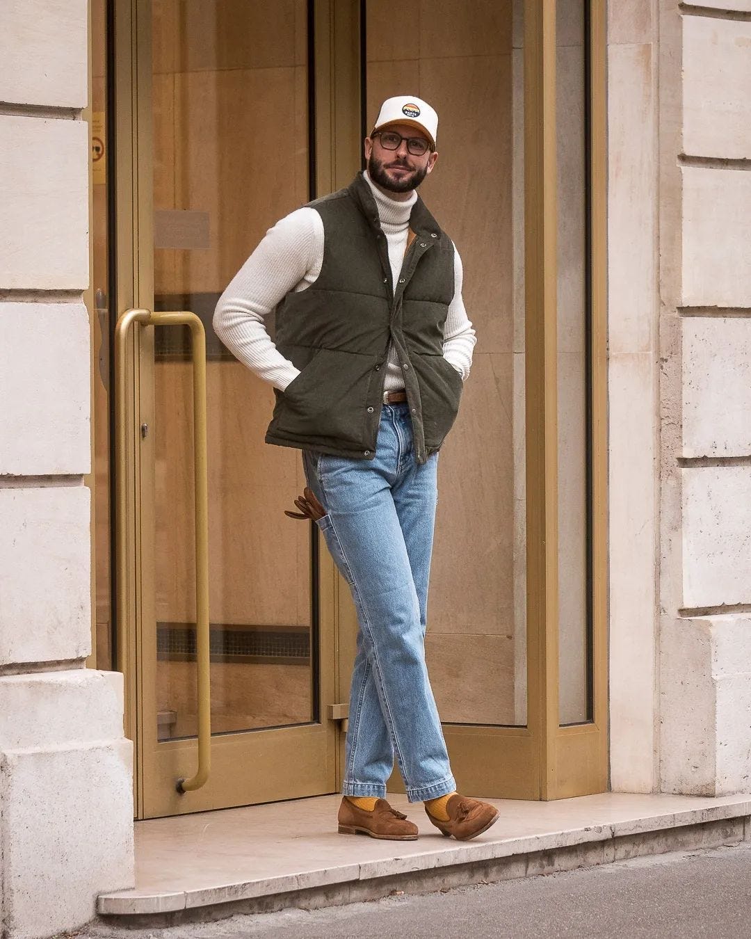stylish man wearing an off-white turtleneck with an olive green vest and light wash jeans with brown suede loafers