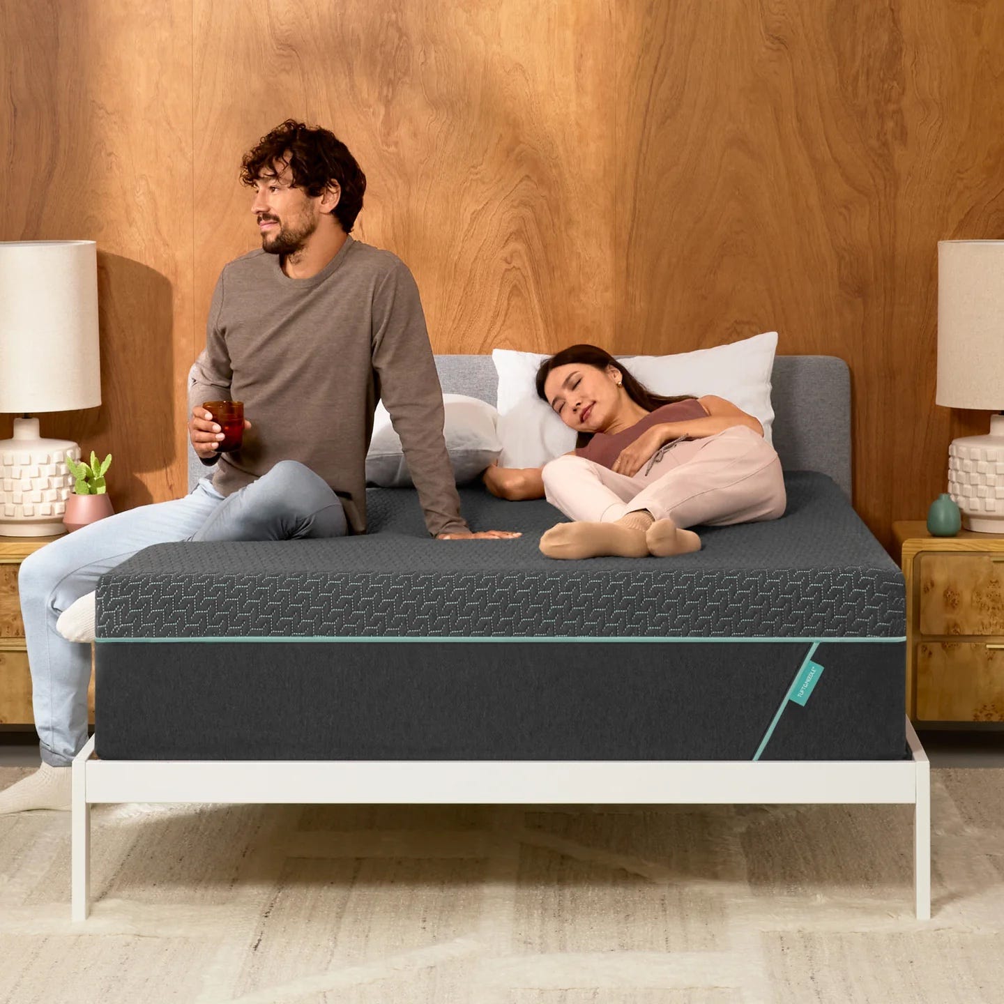 man and woman in pajamas on a bed, the man is drinking a glass of water and the woman is napping